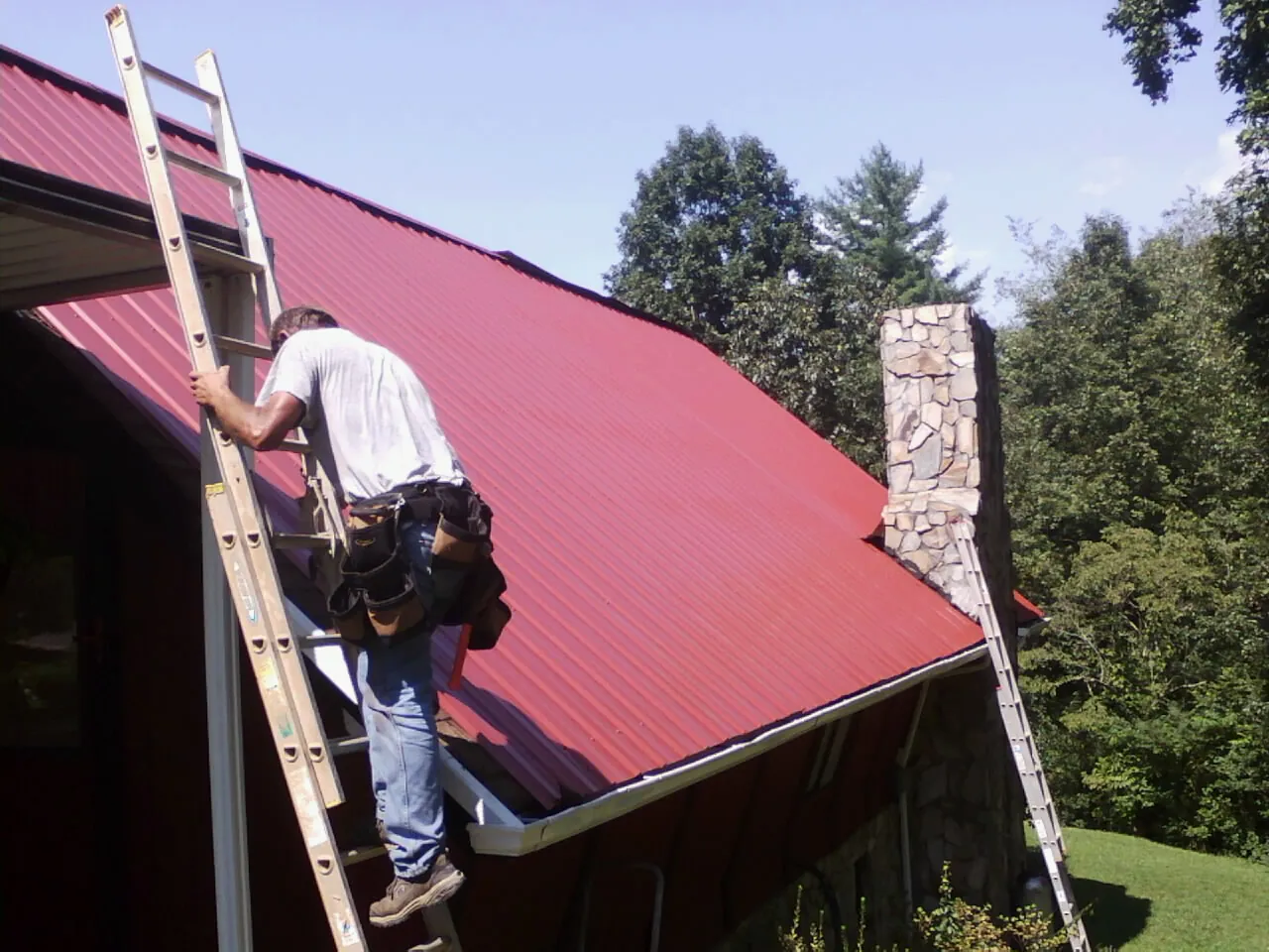 Roofer on a ladder beside a red metal roof and stone chimney — Soffit & Fascia Repair by Roofing Brothers in Claysville